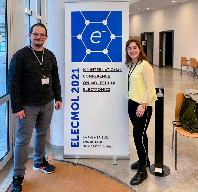 A photograph of Nerea González and Alejandro De Sousa next to a banner of the ELECTROMOL 2021 Conference.